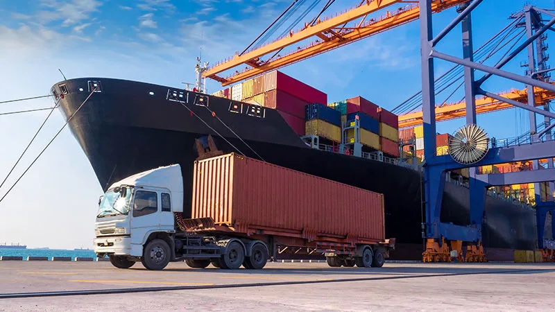 Truck carrying a shipping container beside a cargo ship at a port terminal